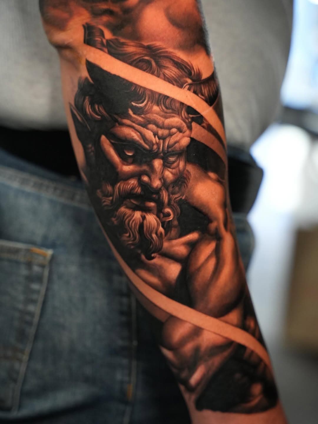 Black and grey realistic Zeus statue tattoo with ribbon detail on forearm by Ink Theory